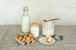 A rustic display of baking ingredients including eggs, milk bottle, and flour on a linen surface.